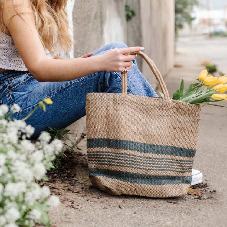 Eco-friendly jute tote bag in slate blue and natural color block design, perfect for groceries, books, beach items, and everyday essentials. The product photo features a hip girl carrying the bag with yellow tulips.